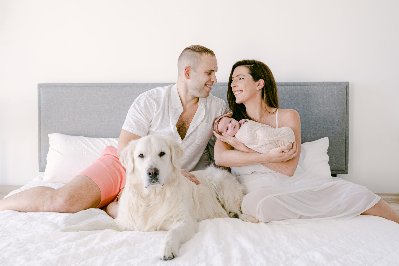 Family smiling at each other on the bed with their newborn baby and their pet