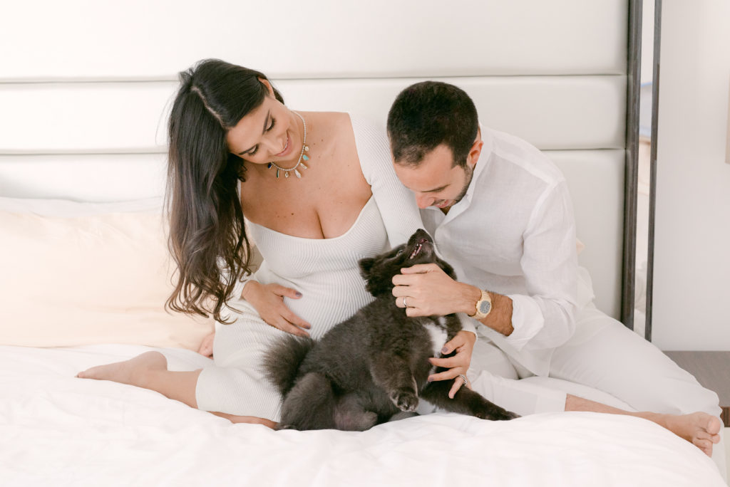 Lifestyle photo of a dad and mom-to-be playing with their pet on the bed in Miami Beach, FL