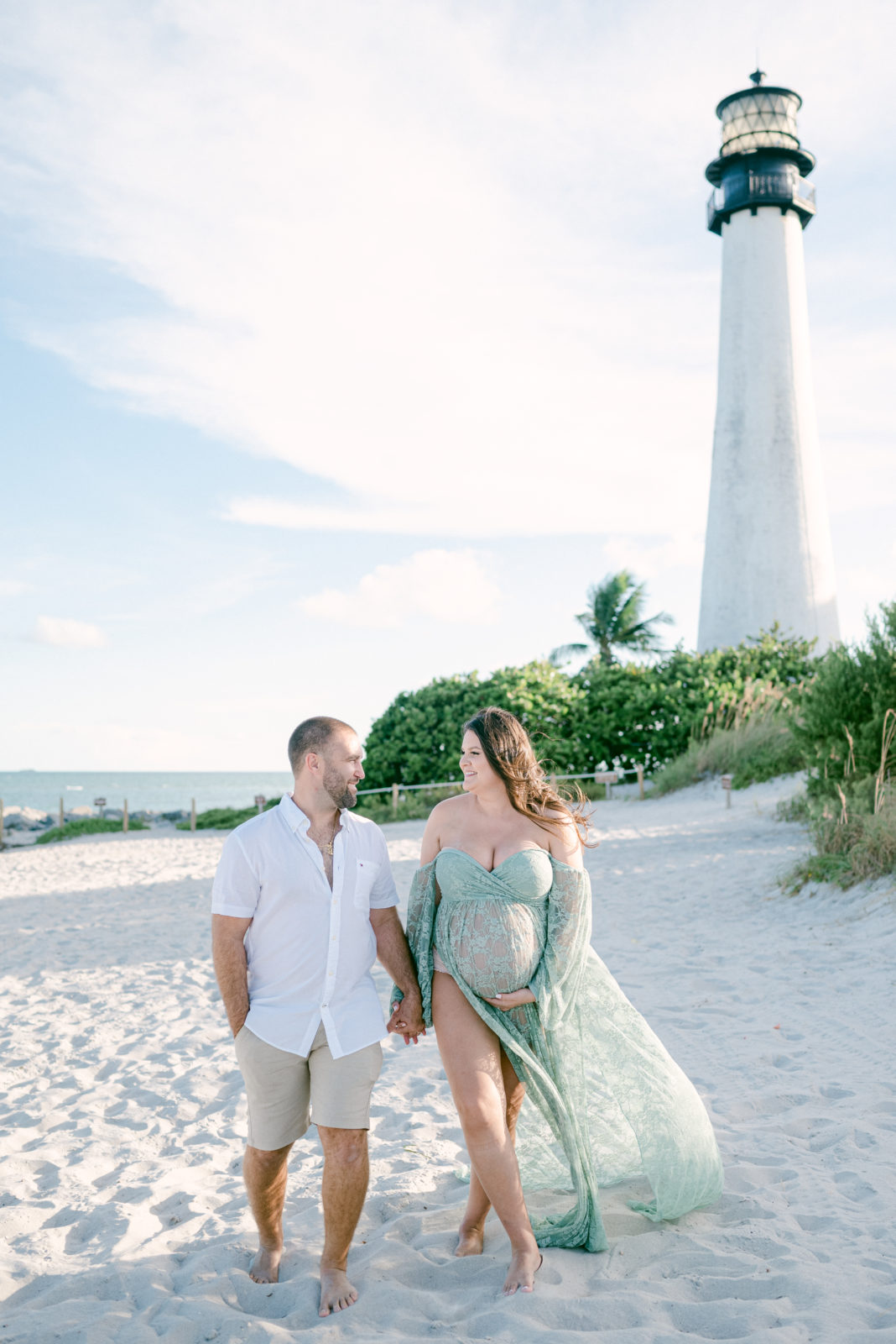 Expecting couple walking on the beach with the lighthouse in the back