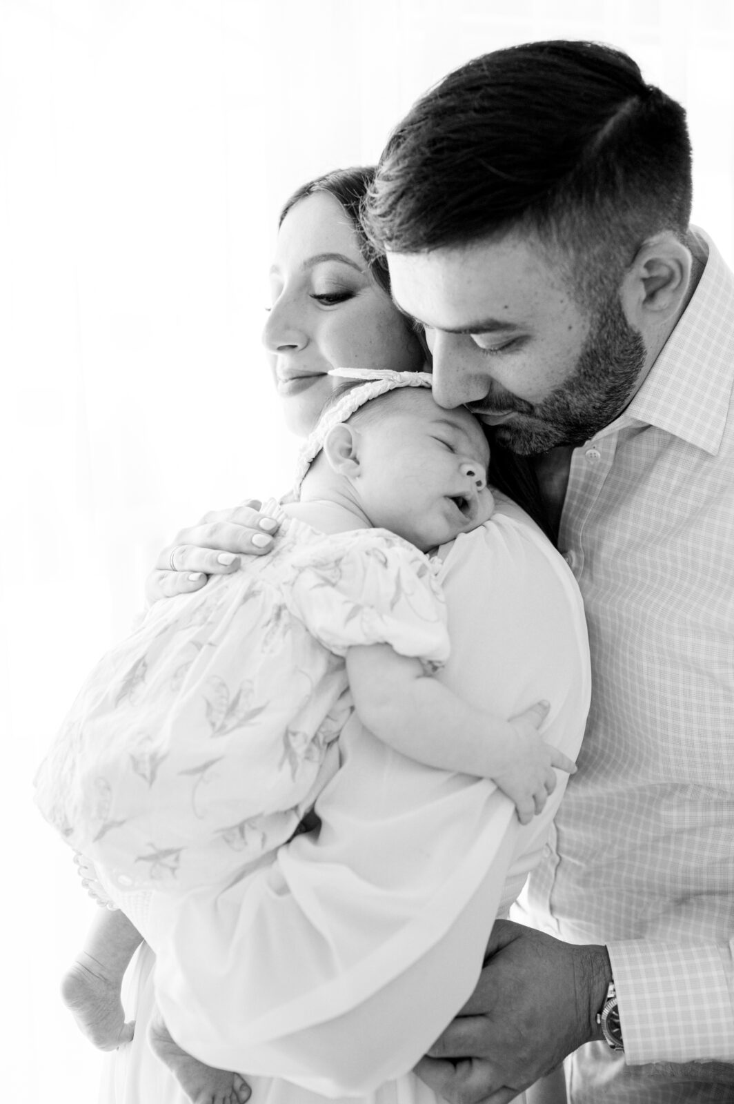 Black and white portrait of new parents with baby girl. Dad is kissing baby on the forehead